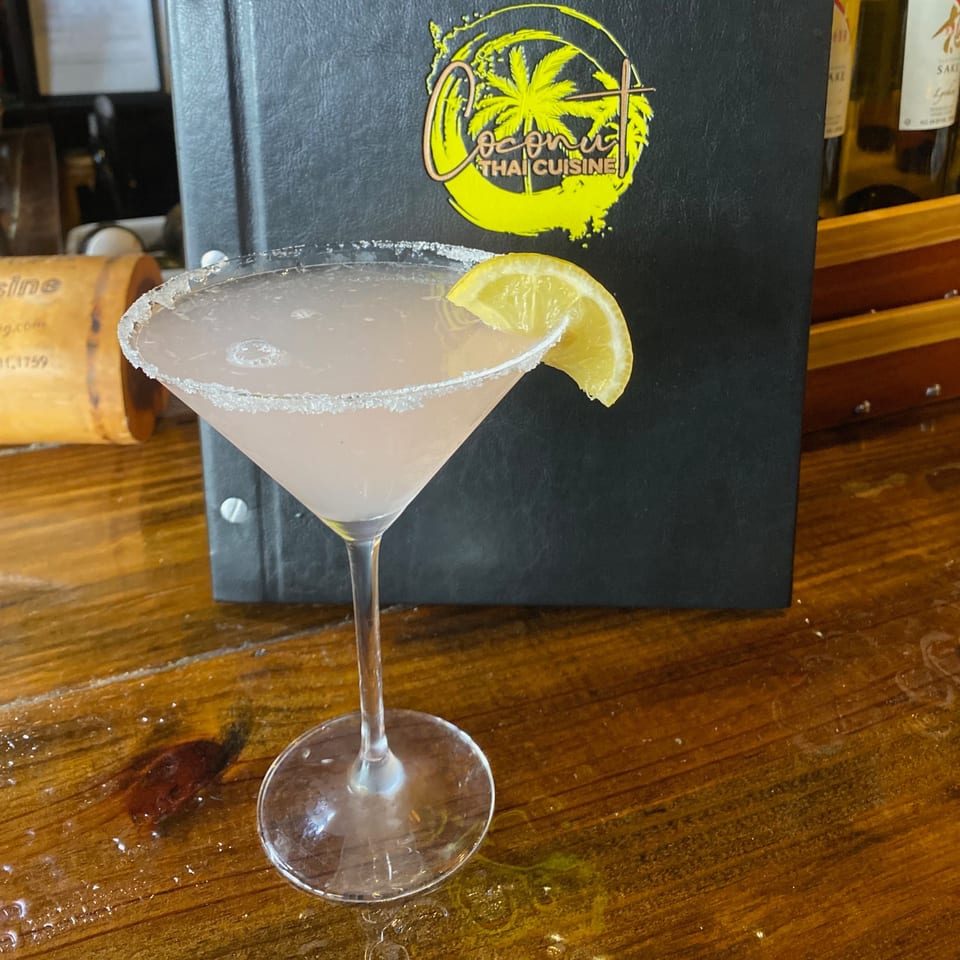 A pink martini with a sugar-rimmed glass and lemon wedge, in front of a Coconut Thai Cuisine menu.