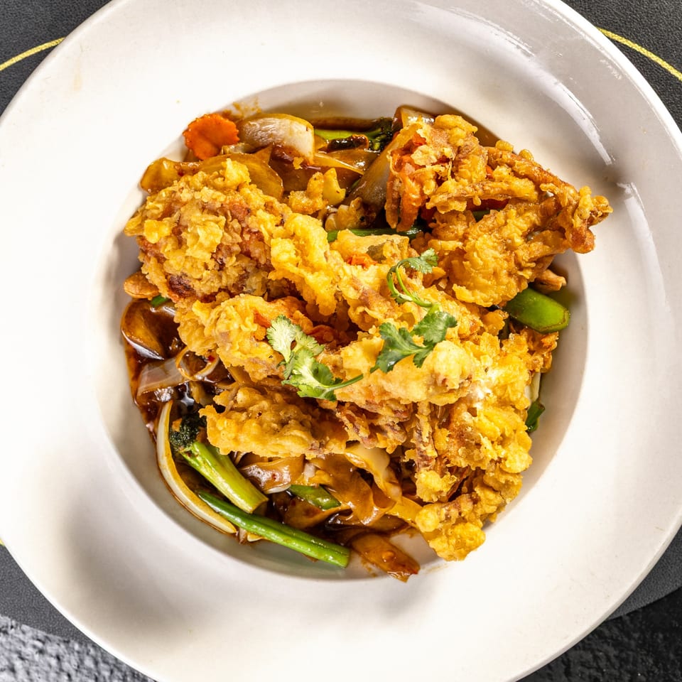 Crispy fried soft-shell crab with wide noodles, vegetables, and dark sauce in a white bowl.