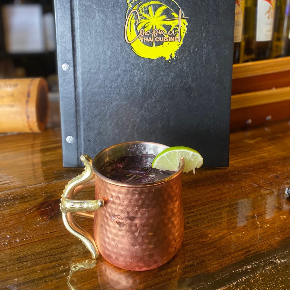 A Moscow Mule in a copper mug with a lime wedge sits on a wooden bar next to a Coconut Thai Cuisine menu.