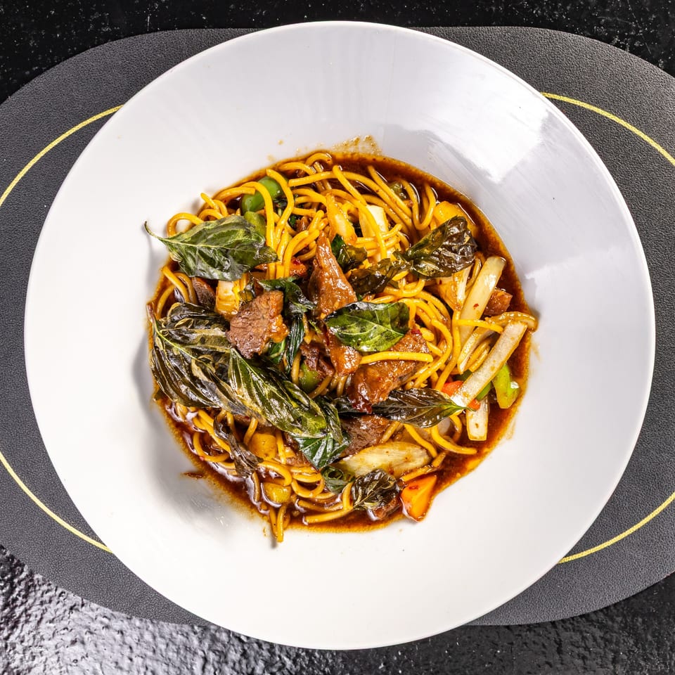 A white plate holds a dish of noodles, beef, vegetables, and basil in a dark sauce.
