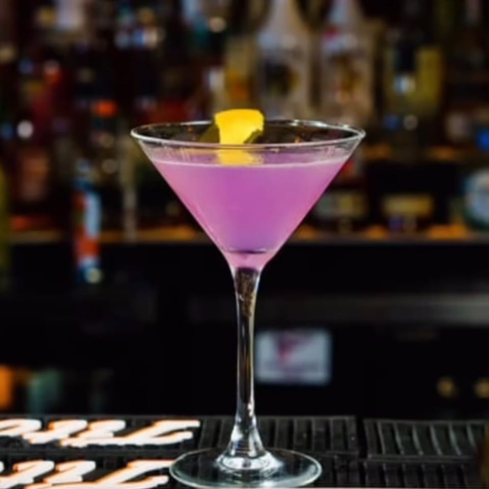 A vibrant purple martini with a lemon twist sits on a bar, with blurred bottles in the background.