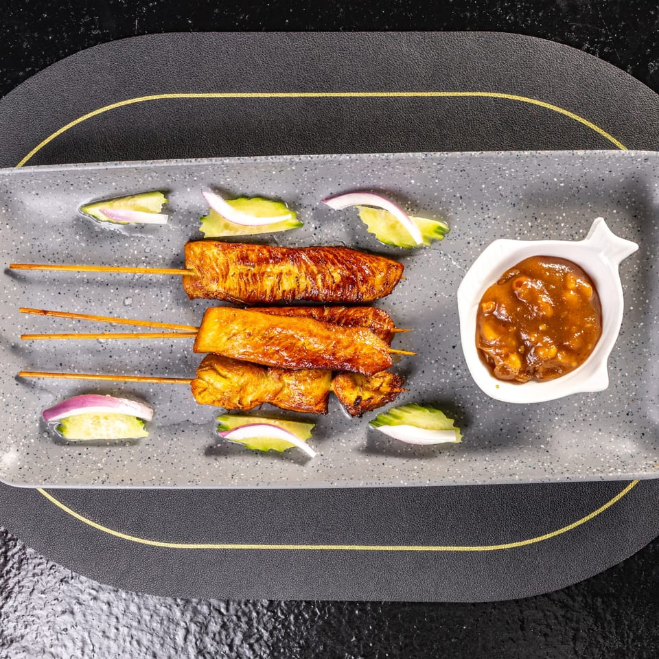 Grilled chicken satay skewers with peanut sauce, cucumber, and red onion on a gray platter.