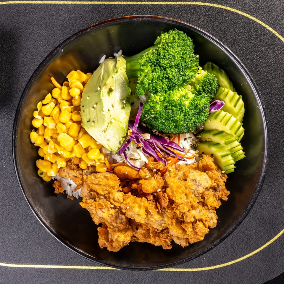 A colorful poke bowl with crispy chicken, corn, avocado, broccoli, cucumber, and purple cabbage.