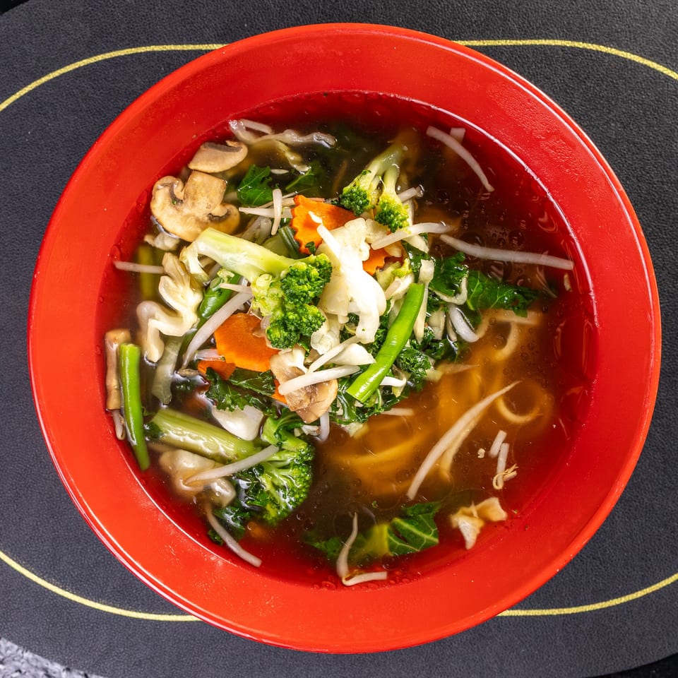 A vibrant vegetable noodle soup in a red bowl, featuring broccoli, carrots, mushrooms, and greens.