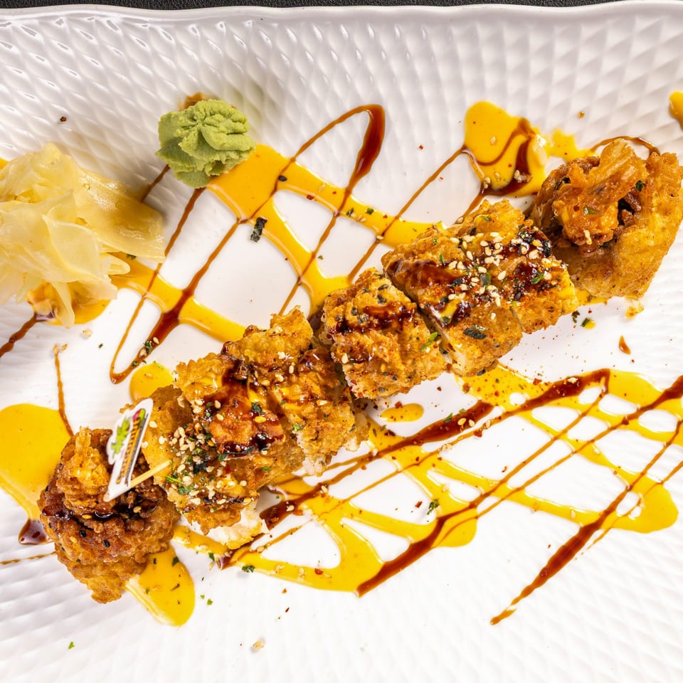 Crispy fried sushi rolls drizzled with yellow and brown sauces, garnished with sesame seeds, wasabi, and ginger.